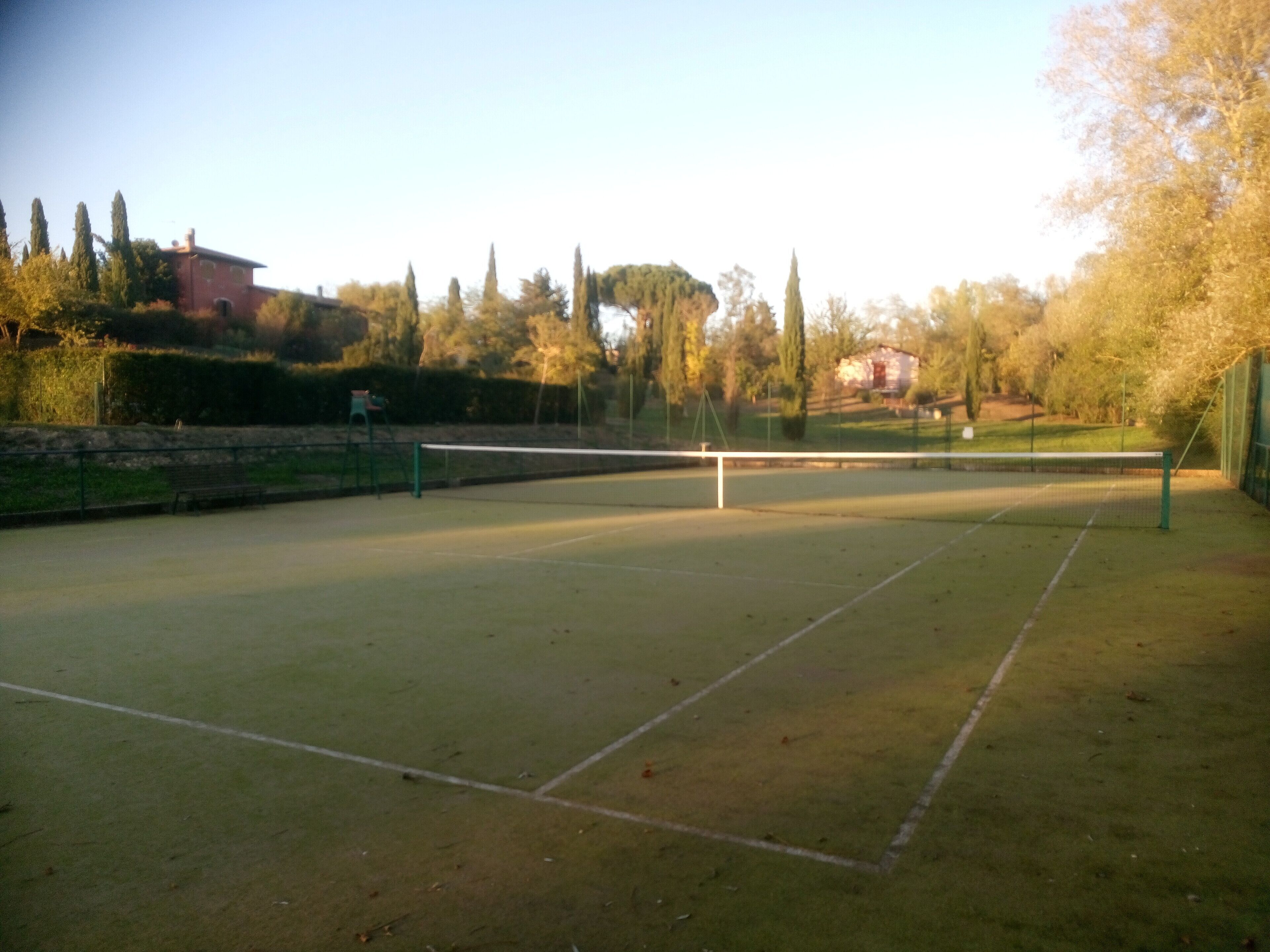 En udendørs tennisbane med net, omgivet af træer og en bygning i baggrunden.