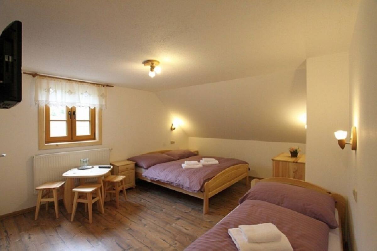 Ein kleines, gemütliches Schlafzimmer mit zwei Betten, einem Holztisch und einem Stuhl. Ein Fenster mit Vorhängen, ein Heizkörper und eine Wandleuchte sind ebenfalls vorhanden.