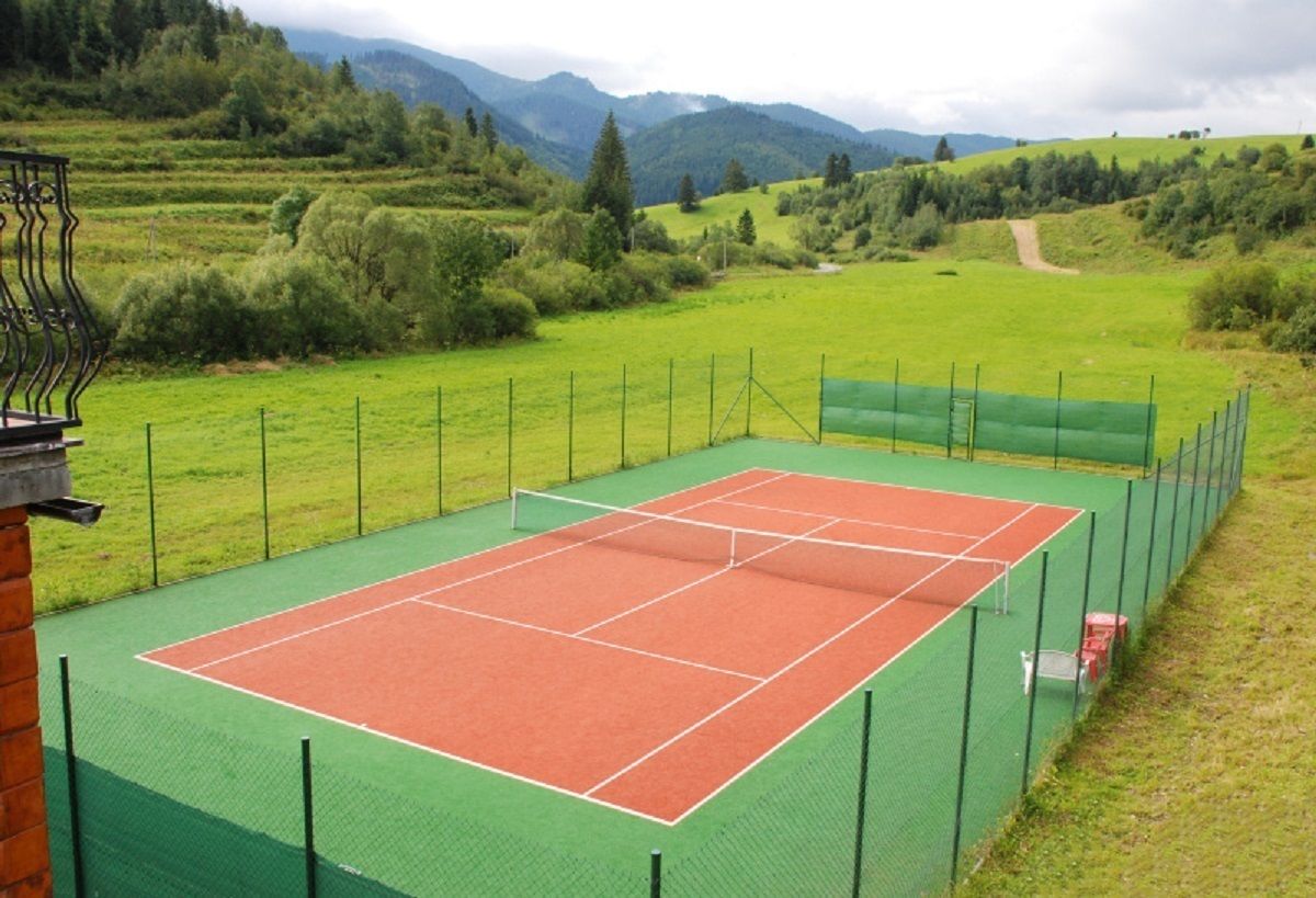 A tennis court with a green fence, surrounded by a grassy field and mountains in the background.