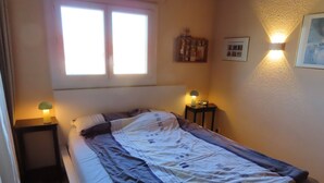 Una cama individual con una colcha blanca y azul, dos lámparas de noche, una ventana con persianas y un estante pequeño con objetos.