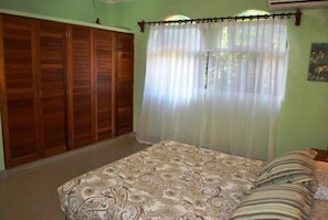 A bedroom with a bed, a wooden wardrobe, and a window with sheer curtains.