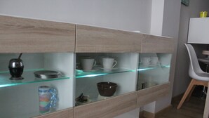 A modern kitchen with wooden cabinets, a glass shelf with kitchenware, and a white chair.