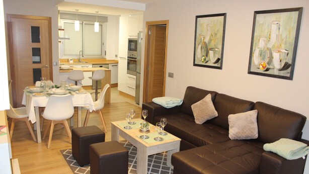 A modern living room with a brown leather sofa, a dining table set for four, and a kitchen in the background.