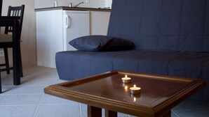 A compact living space with a dark blue sofa, a wooden coffee table with candles, a small kitchenette, and a dining area with a chair. - Villas Lidia - Fengari (Livadi)