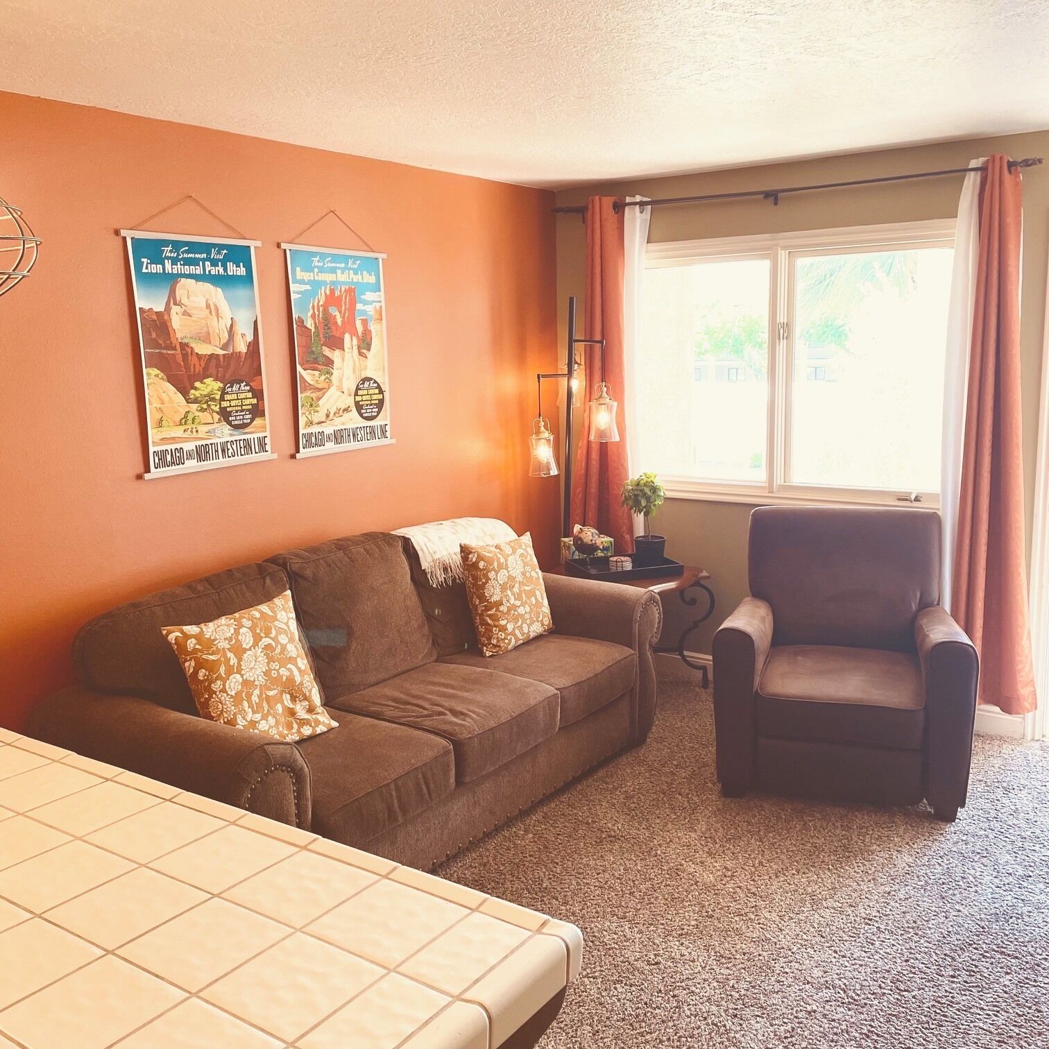 A room with a brown sofa, a chair, a small table with a lamp, and a window with curtains.