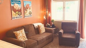 Ein Zimmer mit einem braunen Sofa, einem Sessel, einem kleinen Tisch mit Lampe und einem Fenster mit Vorhängen.