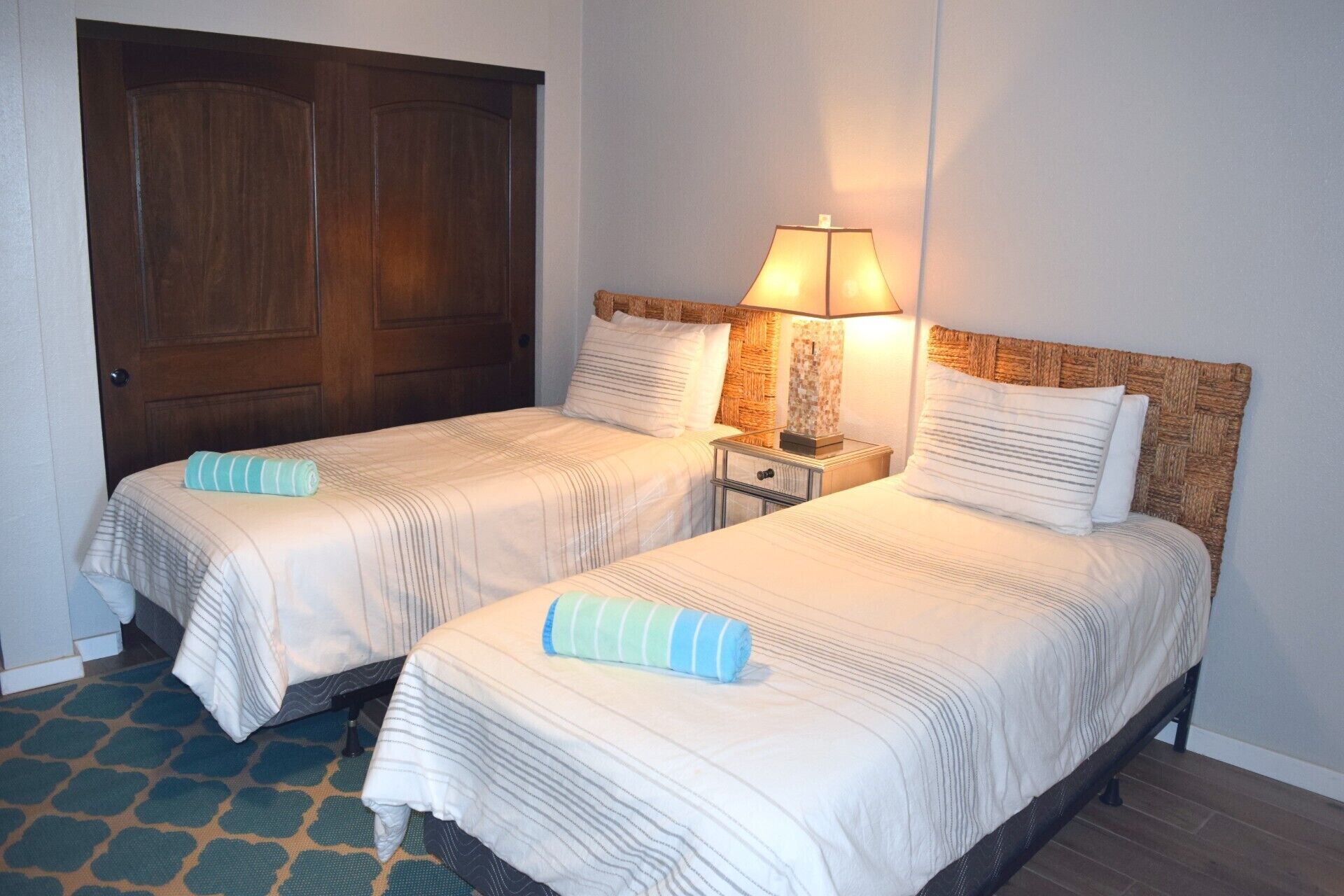 A hotel room with two beds, a wooden door, a nightstand with a lamp, and a patterned floor.