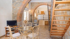 Un coin repas avec une table en verre et des chaises en osier, un téléviseur posé sur un meuble en bois, et un escalier avec une rampe en bois.