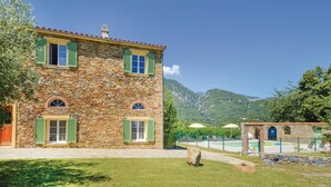 Un bâtiment en pierre avec des volets verts, un chemin en pierre et une chaîne de montagnes en arrière-plan.