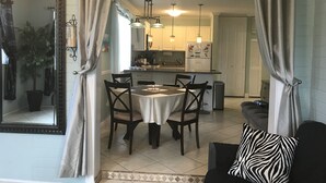 A dining area with a table and chairs, a kitchen in the background, and a large mirror on the left.
