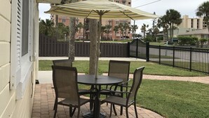 An outdoor patio with a round table and four chairs, a striped umbrella, and a view of a residential area with palm trees and a fence.