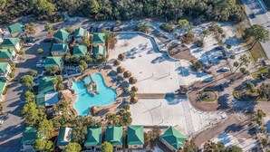 Vista aerea di un resort con piscina centrale, diversi edifici con tetti verdi e una spiaggia sabbiosa.