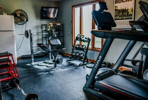 A home gym with a treadmill, weight bench, kettlebells, and a TV.