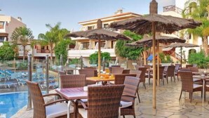 Un espace aménagé au bord de la piscine, avec des fauteuils et des tables en osier, une piscine et une vue sur un immeuble doté d’un balcon.