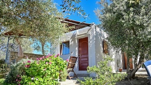 A small white building with a red door and windows, surrounded by greenery and a chair outside. - The Papas House on the Hill (Kamilari)