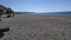 A beach with a thatched-roof structure, a few people, and a clear view of the sea and distant hills. - The Papas House on the Hill (Kamilari)