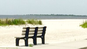 A bench on a sandy beach with a clear view of the water and a distant shoreline. - Peaceful Gulf Getaway - Close to Old Town Bay St Louis (Bay St Louis)