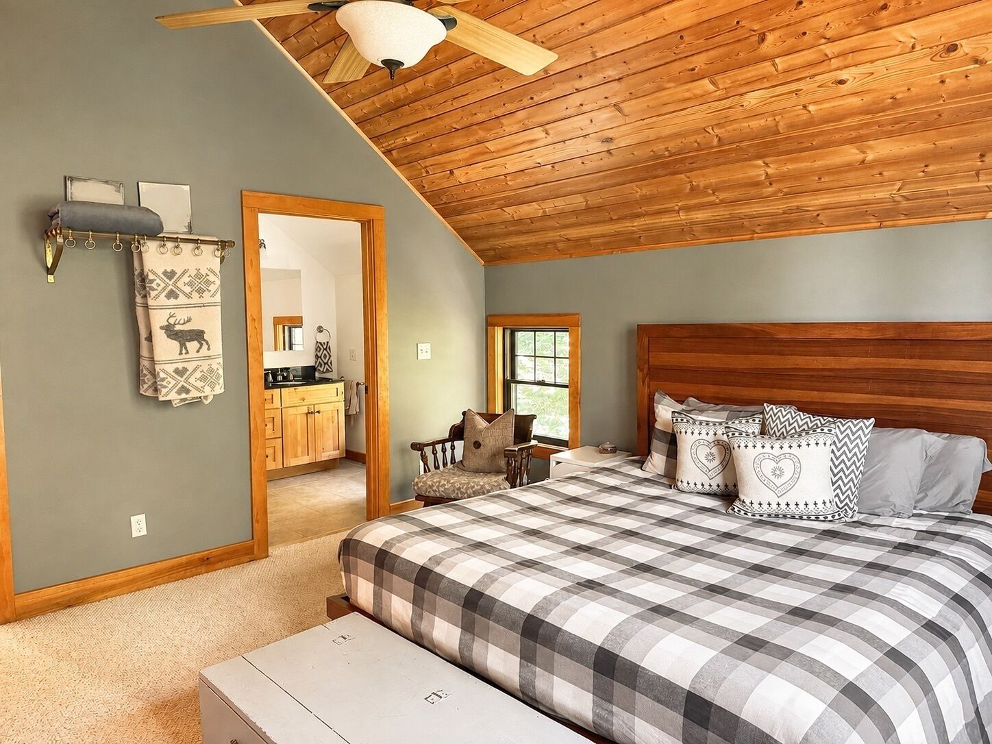 Une chambre avec un plafond en bois, un lit recouvert d’une courtepointe à carreaux et une vue sur la cuisine par une porte ouverte.