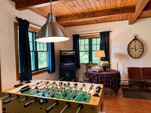 A room with a foosball table, a table with a lamp and flowers, a clock on the wall, and a window with curtains.