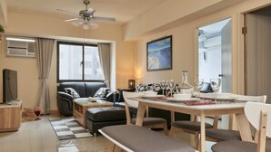 Una sala de estar moderna con un sofá, una mesa de comedor y un banco. Hay un ventilador de techo, una ventana con cortinas y un cuadro en la pared.