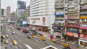Una concurrida calle urbana con varios taxis amarillos, un edificio Sogo imponente y diversos comercios.
