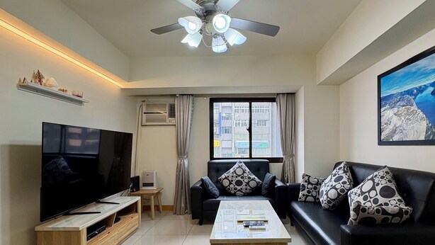 Una sala de estar moderna con un televisor de pantalla plana, un ventilador de techo, un sofá con cojines estampados, una mesa de centro y una ventana con cortinas.