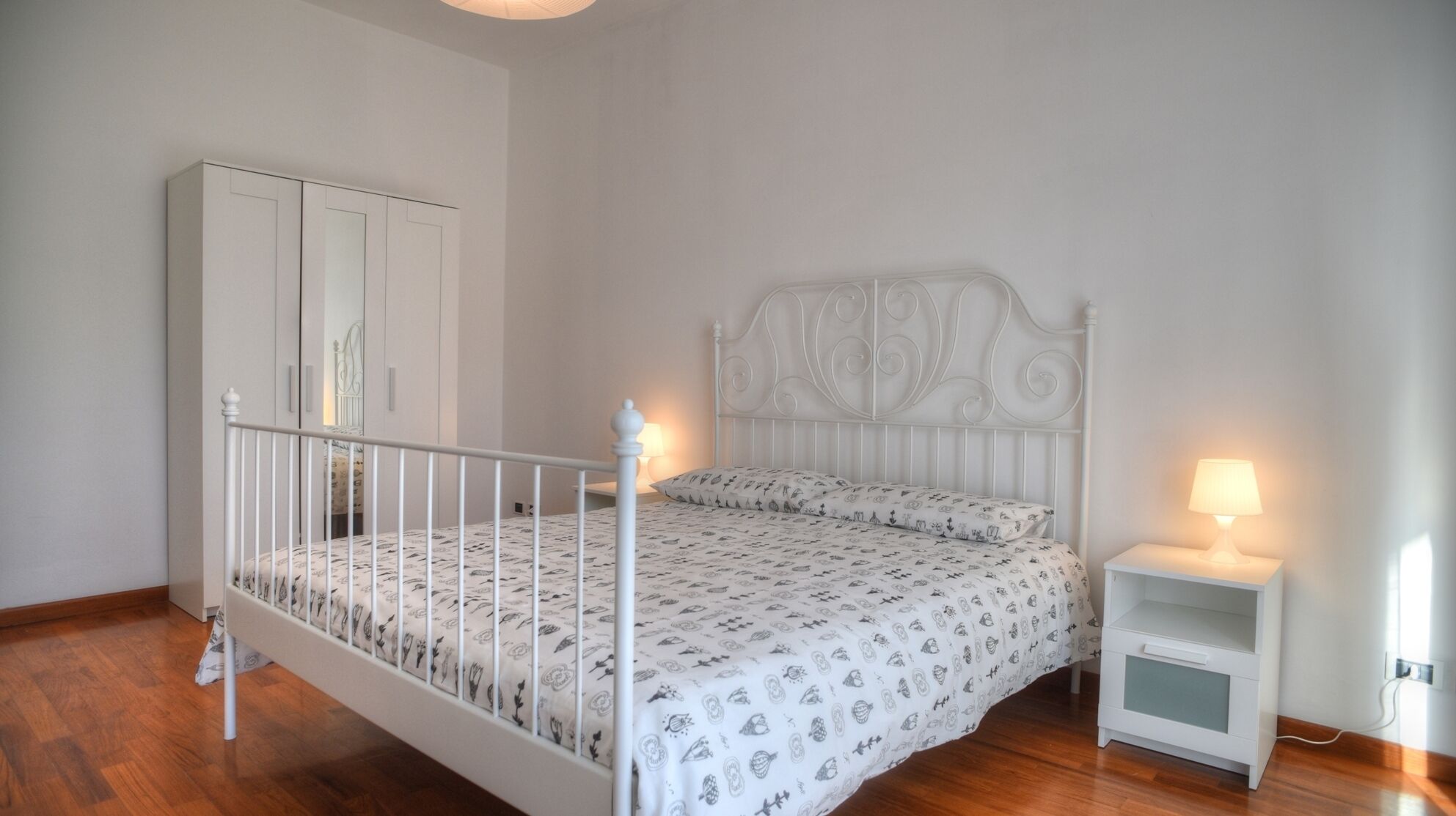 A bedroom with a white metal bed frame, a white nightstand with a lamp, and a wardrobe with sliding doors.