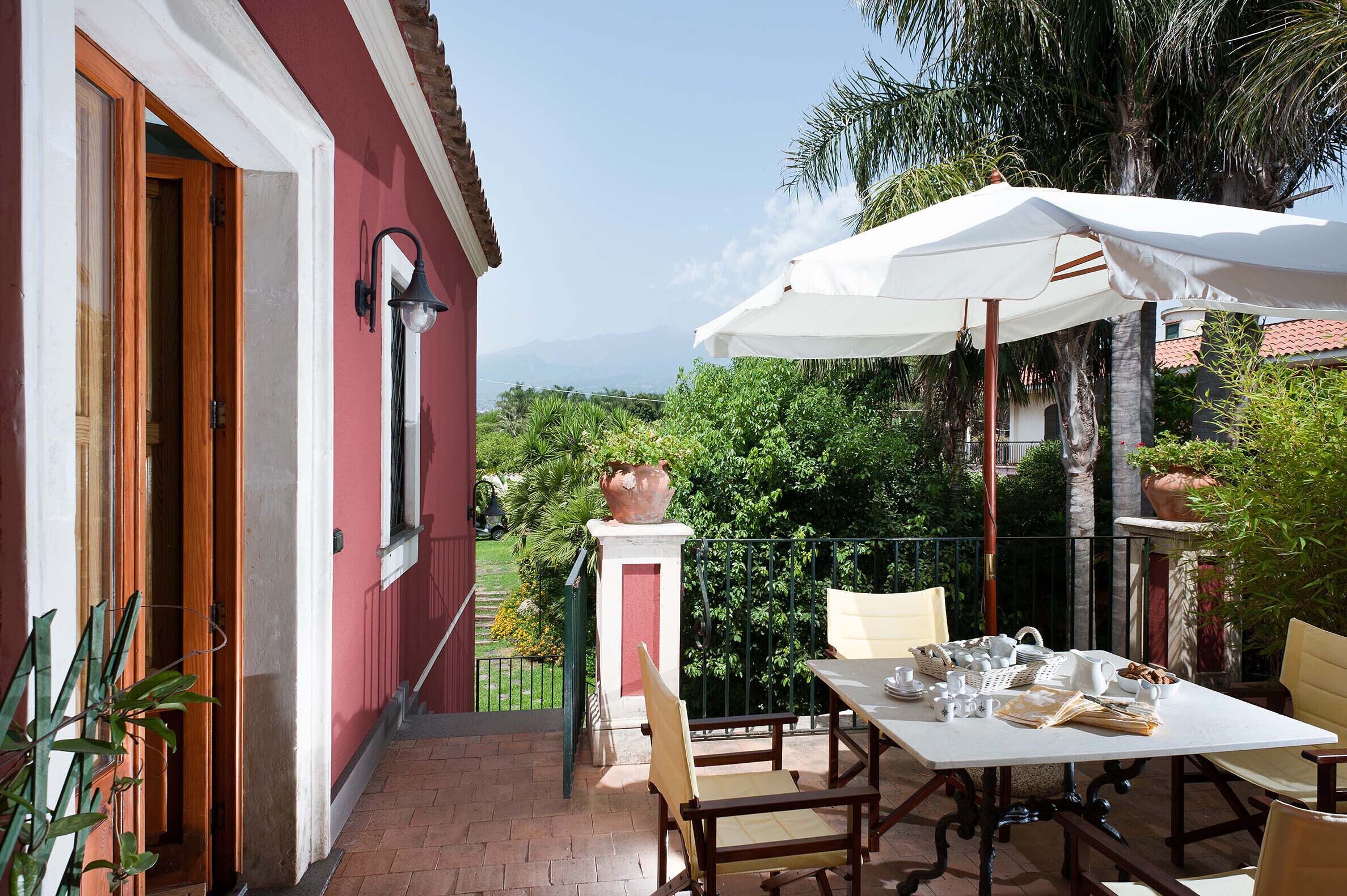 A patio with a table and chairs, a white umbrella, and a view of greenery and mountains.