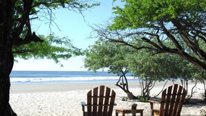 Eine Strandansicht mit zwei hölzernen Adirondack-Stühlen und einem kleinen Tisch unter einem Baum, mit Blick auf den Ozean.
