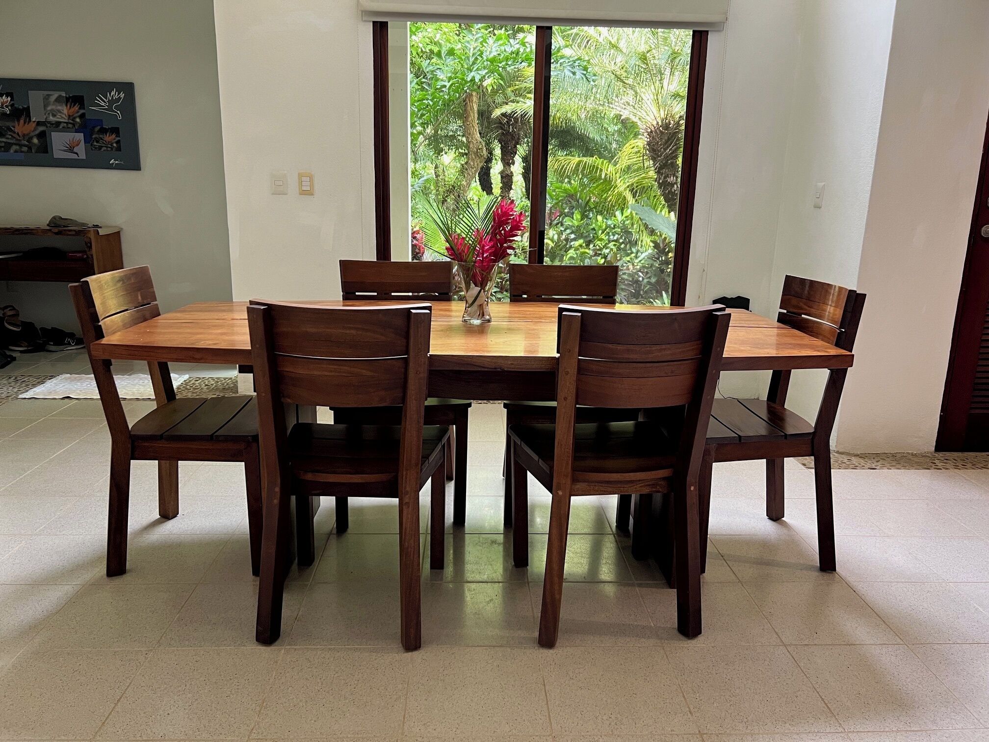 A dining area with a wooden table and chairs, a vase with flowers, and a view of greenery through a window.