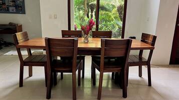 A dining area with a wooden table and chairs, a vase with flowers, and a view of greenery through a window.