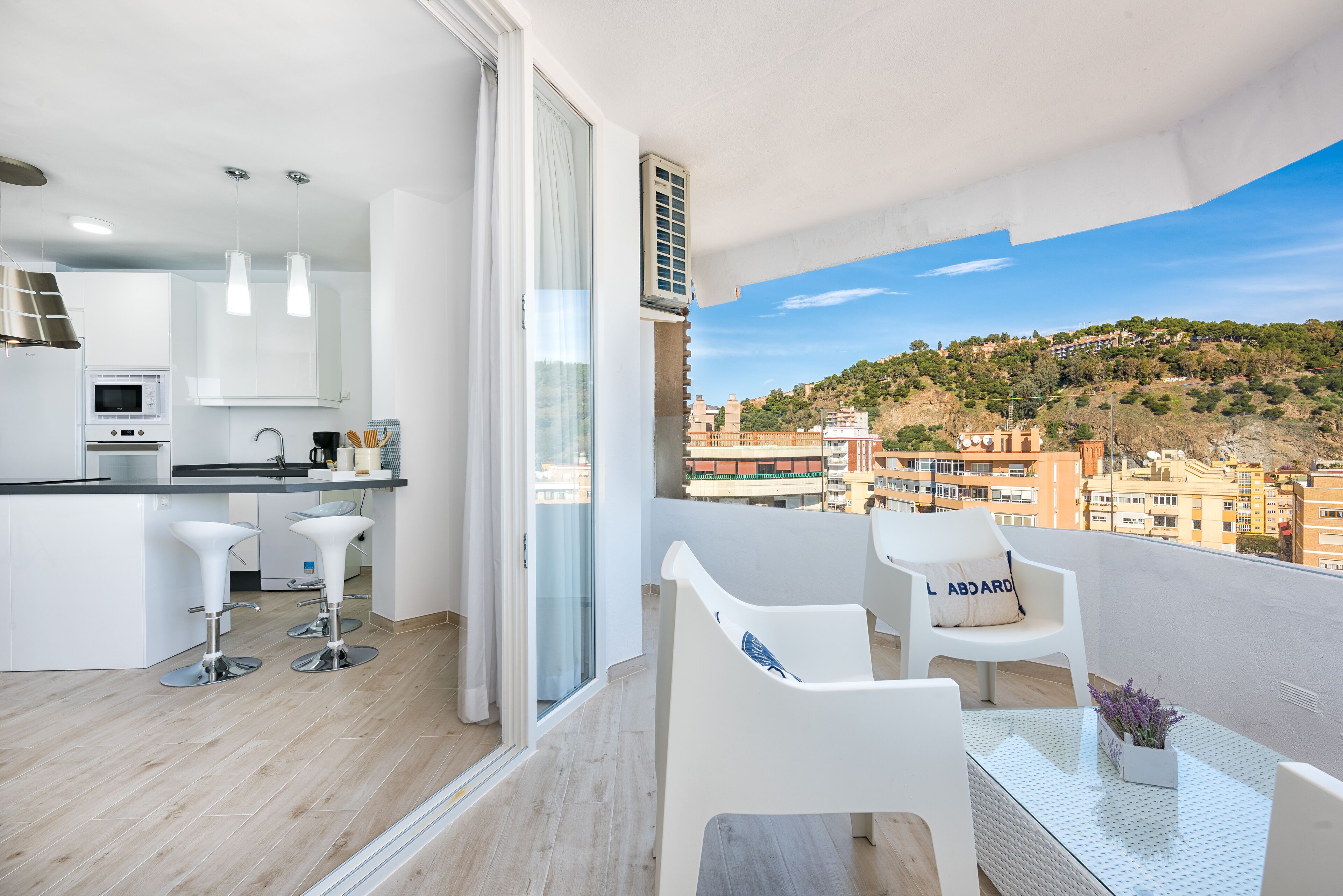 A balcony with a view of a hillside, a kitchen with modern appliances, and white bar stools.