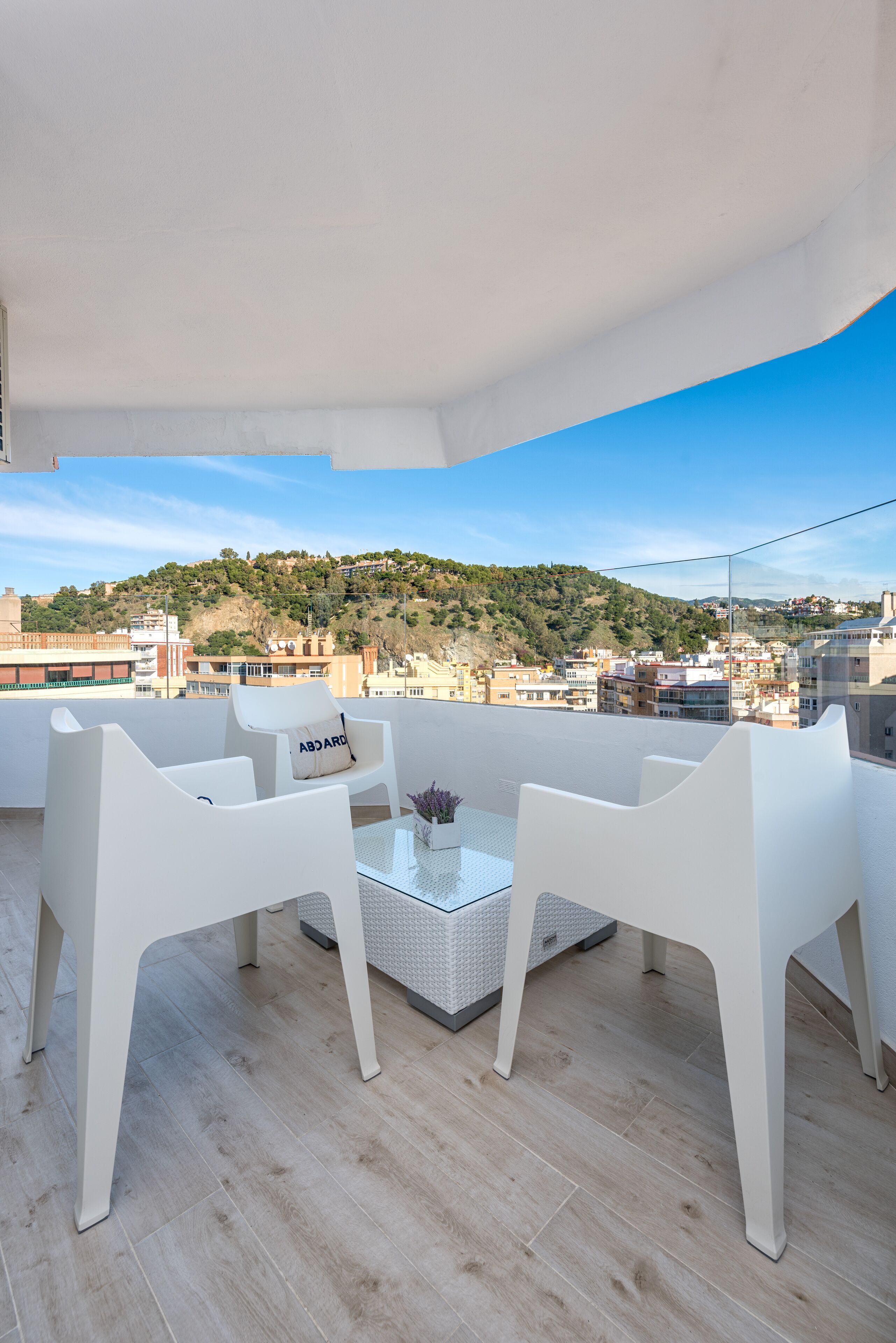 Balcón con sillas blancas y una mesa de vidrio, con vista a un paisaje urbano con edificios y colinas.