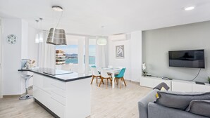 Un appartement moderne comprenant une cuisine, un coin repas et un salon, et doté d’une grande fenêtre offrant une vue sur la mer.