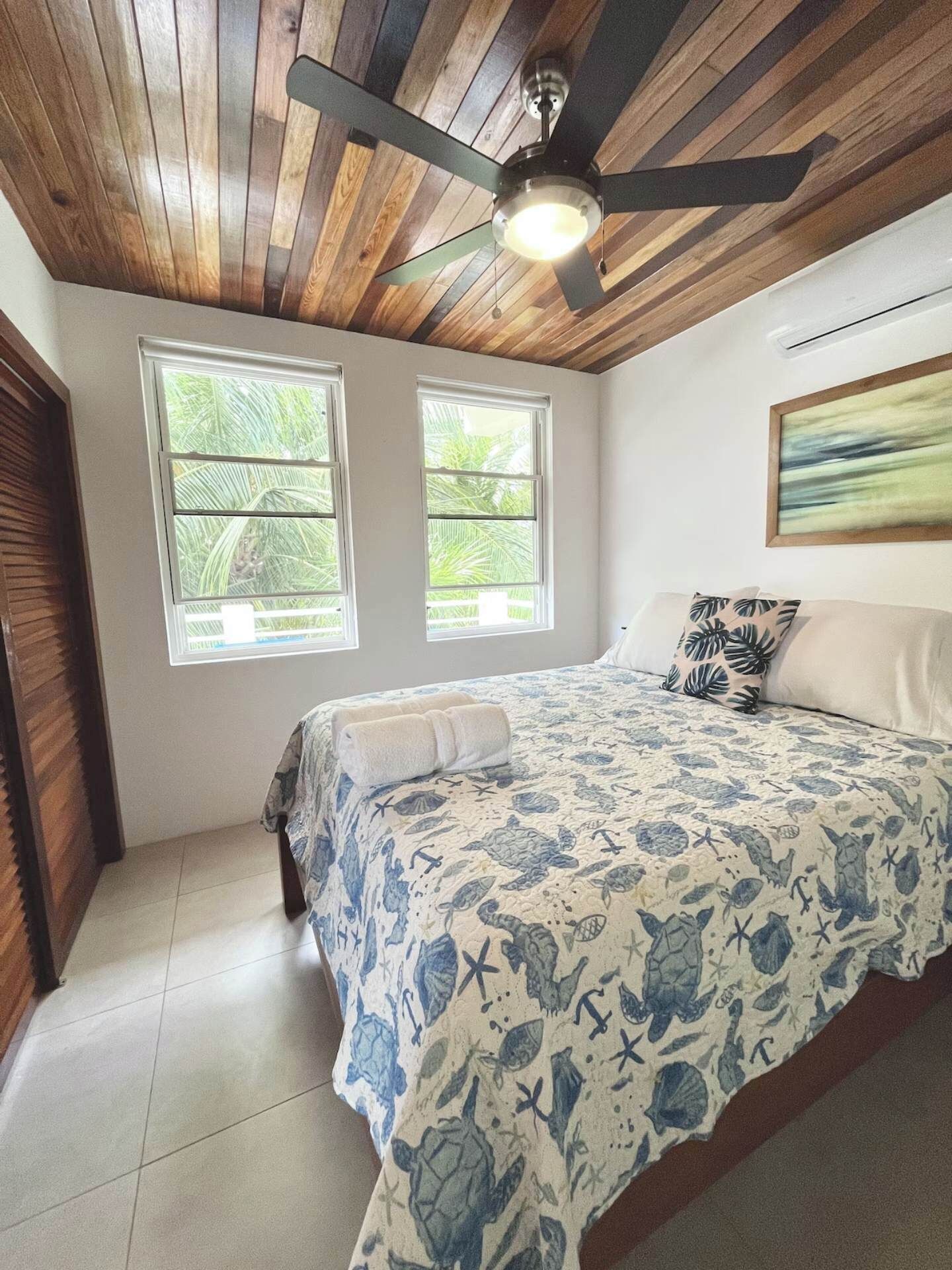 Un dormitorio con techo de madera, ventilador de techo, dos ventanas que dan a vistas de vegetación, una cama con colcha náutica azul y blanca, y una sola almohada.