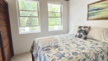 Un dormitorio con techo de madera, ventilador de techo, dos ventanas que dan a vistas de vegetación, una cama con colcha náutica azul y blanca, y una sola almohada.
