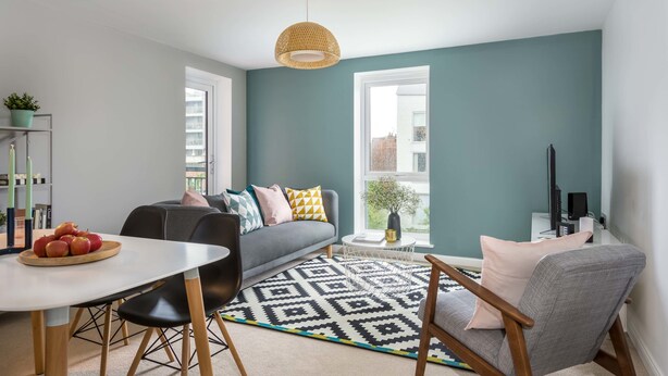 Una sala de estar moderna con un sofá gris, una mesa de comedor con manzanas, una alfombra estampada y un ventanal grande.