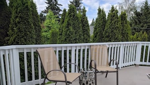 A deck with two chairs and a table, surrounded by a white railing and tall evergreen trees.