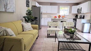 A modern living room with a yellow sofa, two patterned chairs, a glass coffee table, and a kitchen in the background.