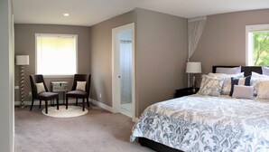 A bedroom with a large bed, two bedside tables with lamps, a chair, a small table, and a window with curtains.