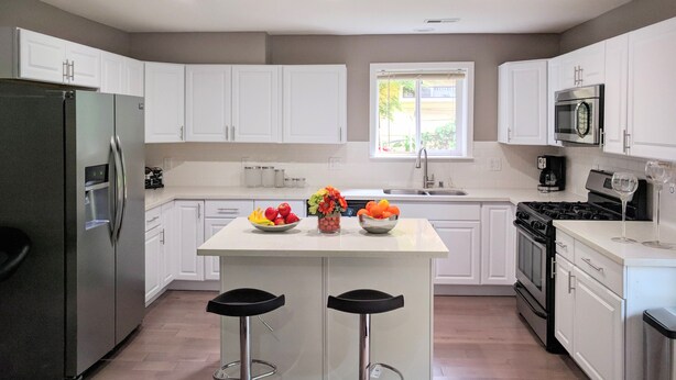 A modern kitchen with white cabinets, a stainless steel refrigerator, a gas stove, and two bar stools.