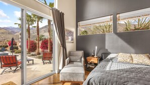 A bedroom with a large sliding glass door leading to an outdoor patio, a bed with a grey quilt, a grey armchair, and a wooden nightstand with a lamp. - Luxury Palm Springs Retreat. Pool, Spa & Stunning Mountain Views (Palm Springs)