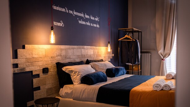 Un dormitorio moderno con un cabecero de ladrillo, una lámpara colgante, un perchero de madera y toallas ordenadamente dobladas sobre la cama.
