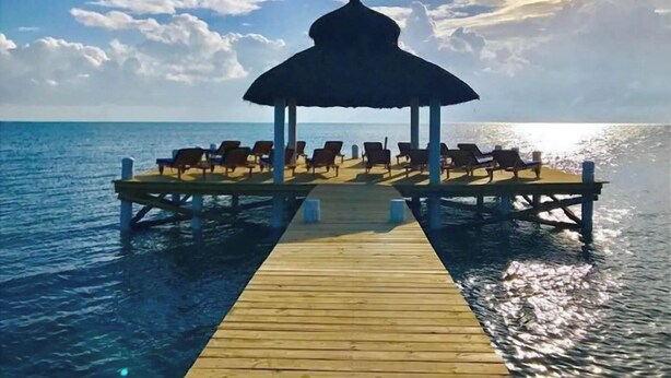 Un muelle de madera que conduce a una playa con sillas de descanso y una estructura de techo de paja.