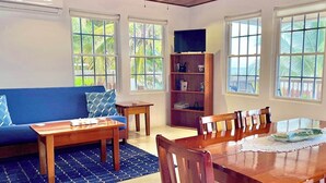 Amplia sala de estar con un sofá azul, una mesa de centro de madera y una mesa de comedor con sillas. Hay una estantería y un ventilador de techo.