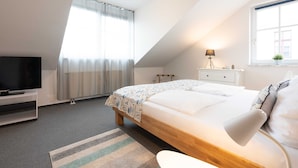 Un dormitorio con una cama, televisión, mesita de noche, lámpara y ventana con cortinas.