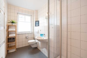 Un bagno con il water, un lavandino e una doccia. C'è una finestra e uno scaffale con asciugamani e una pianta.