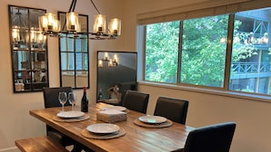 A dining area with a wooden table, chairs, and a bench. There are plates, glasses, and a bottle on the table. A chandelier hangs above, and a large window provides a view of greenery outside.