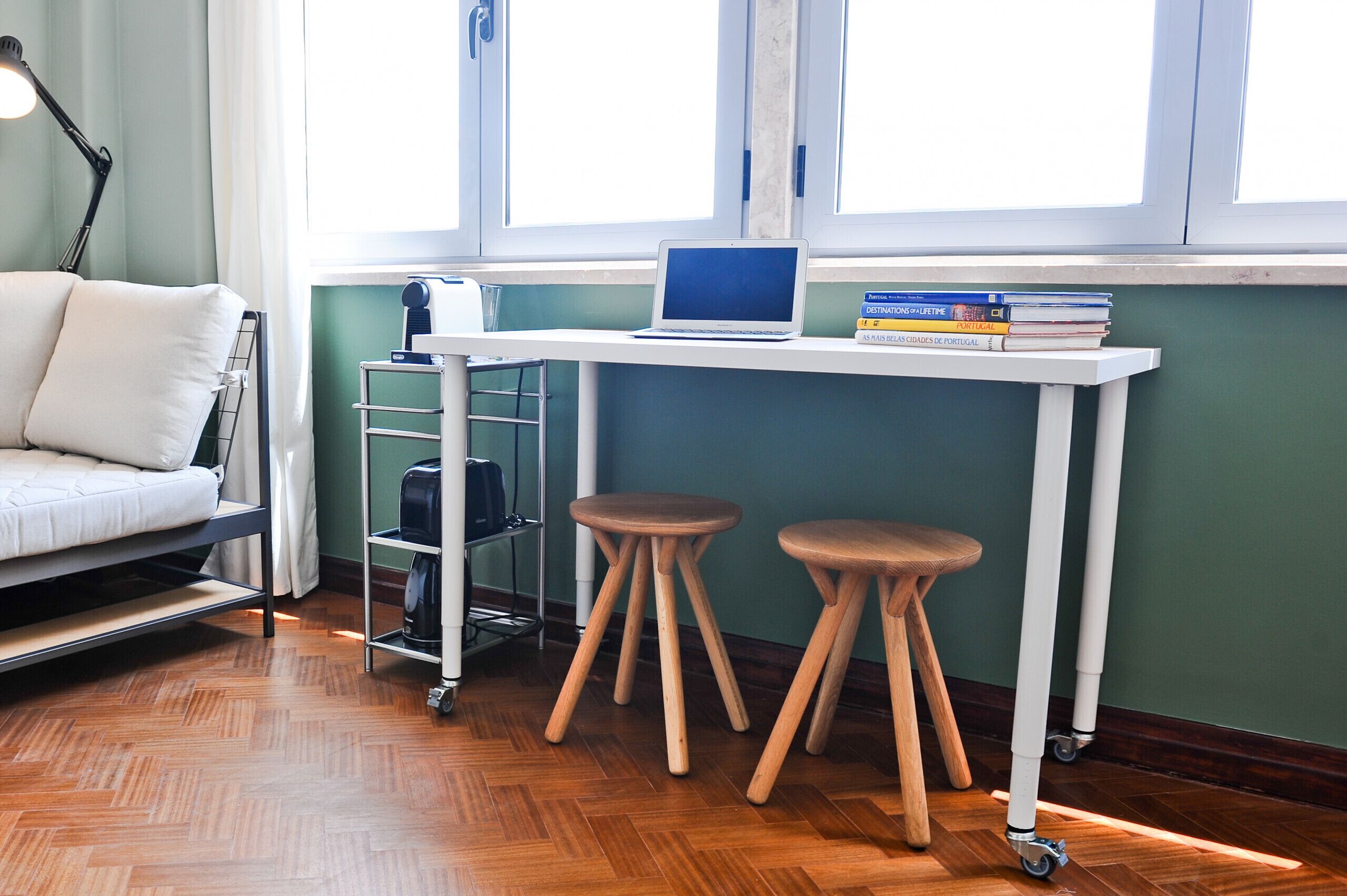 Une pièce de taille modeste comprenant un lit, un bureau avec un ordinateur portable et des livres, une machine à café et une fenêtre avec des rideaux.