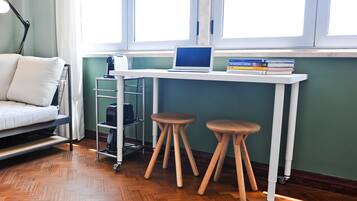 Une pièce de taille modeste comprenant un lit, un bureau avec un ordinateur portable et des livres, une machine à café et une fenêtre avec des rideaux.